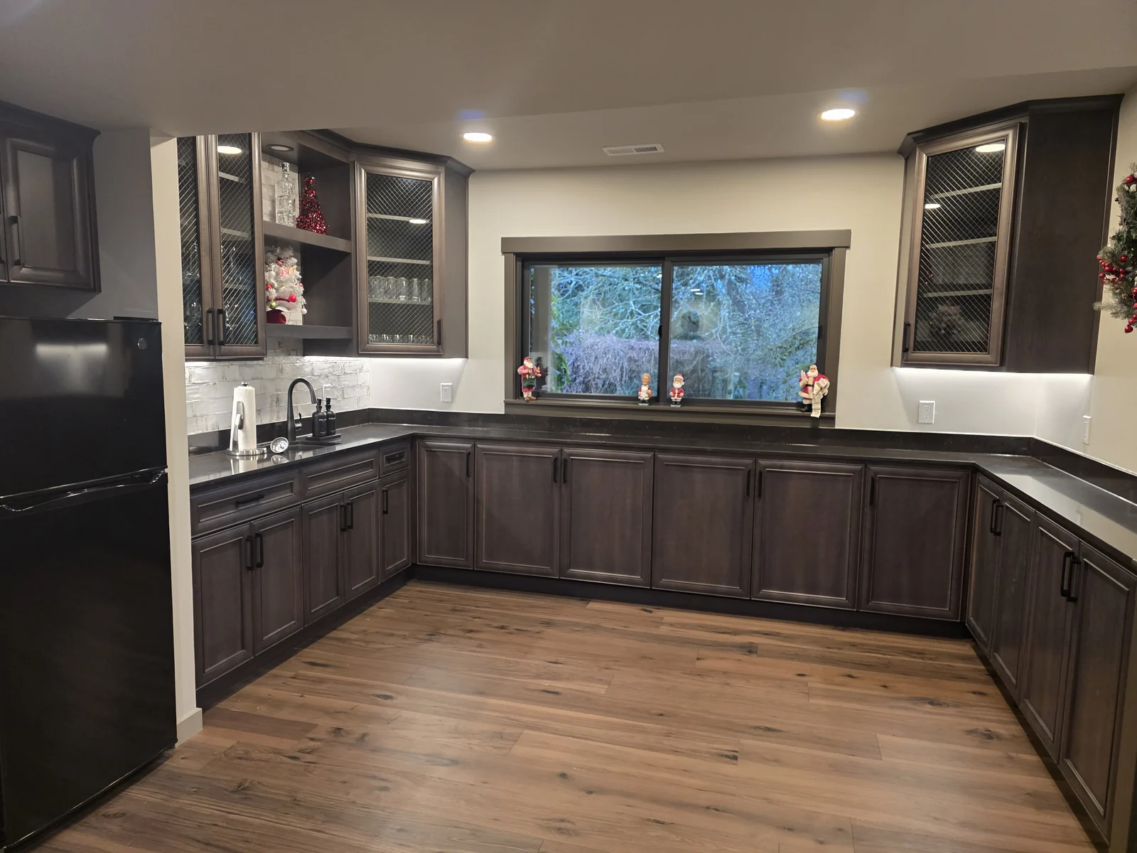 Custom kitchenette conversion in Kent, WA, with espresso-stained Craftsman-style cabinetry and trim, chicken-wire-embedded glass cabinet doors, a custom Milgard Ultra window, stone countertops, a tile backsplash, custom sink installation, and hardwood flooring.