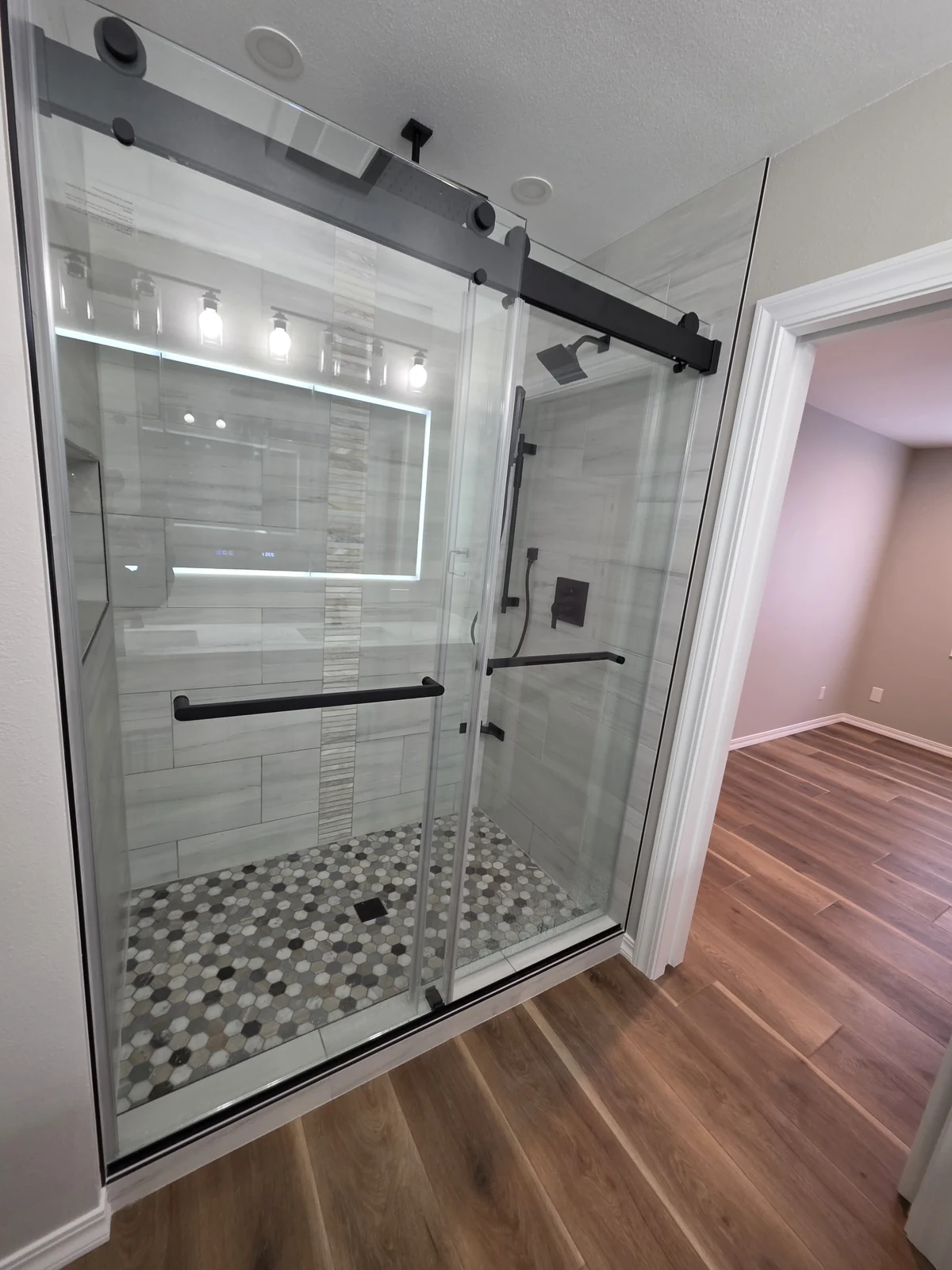 Custom bathroom remodel in Bellevue, WA, featuring a frameless glass shower enclosure with matte-black sliding hardware, tiled shower walls, a built-in recessed niche, a mosaic shower floor, coordinated black Delta fixtures, luxury vinyl plank flooring, and custom open storage.